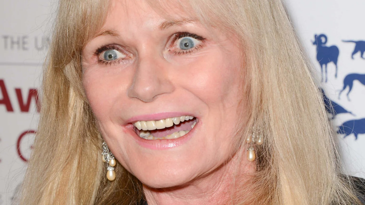Close-up of a blonde woman with wide blue eyes, an open-mouthed smile showing teeth, and pearl drop earrings, against a blurred background with text and animal silhouettes.