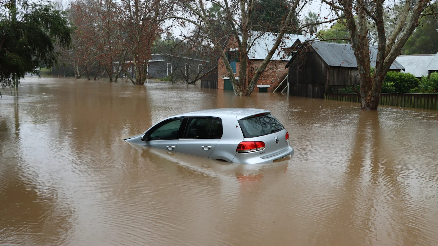 A silver car is partially submerged in murky brown floodwater on a street, with residential homes and bare trees also standing in the water under an overcast sky.