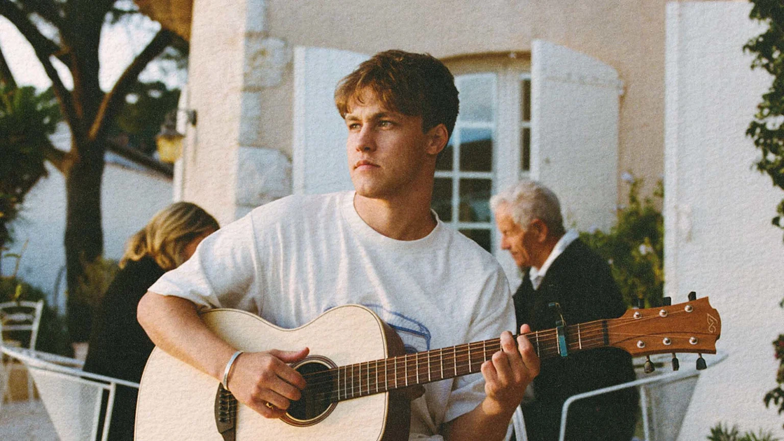 Max Allais, a young man, plays an acoustic guitar outdoors, looking left, with two people and a white building visible in the grainy background.