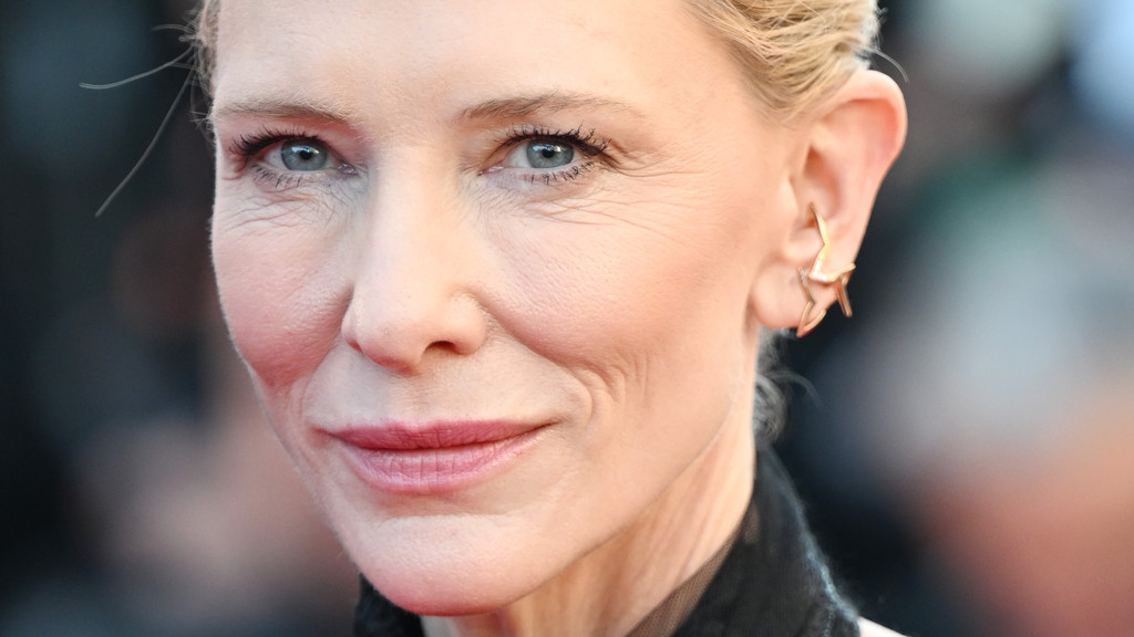 Close-up portrait of Cate Blanchett with blue eyes, light makeup, and a gold star ear cuff on her right ear, with visible wrinkles around her eyes and mouth.