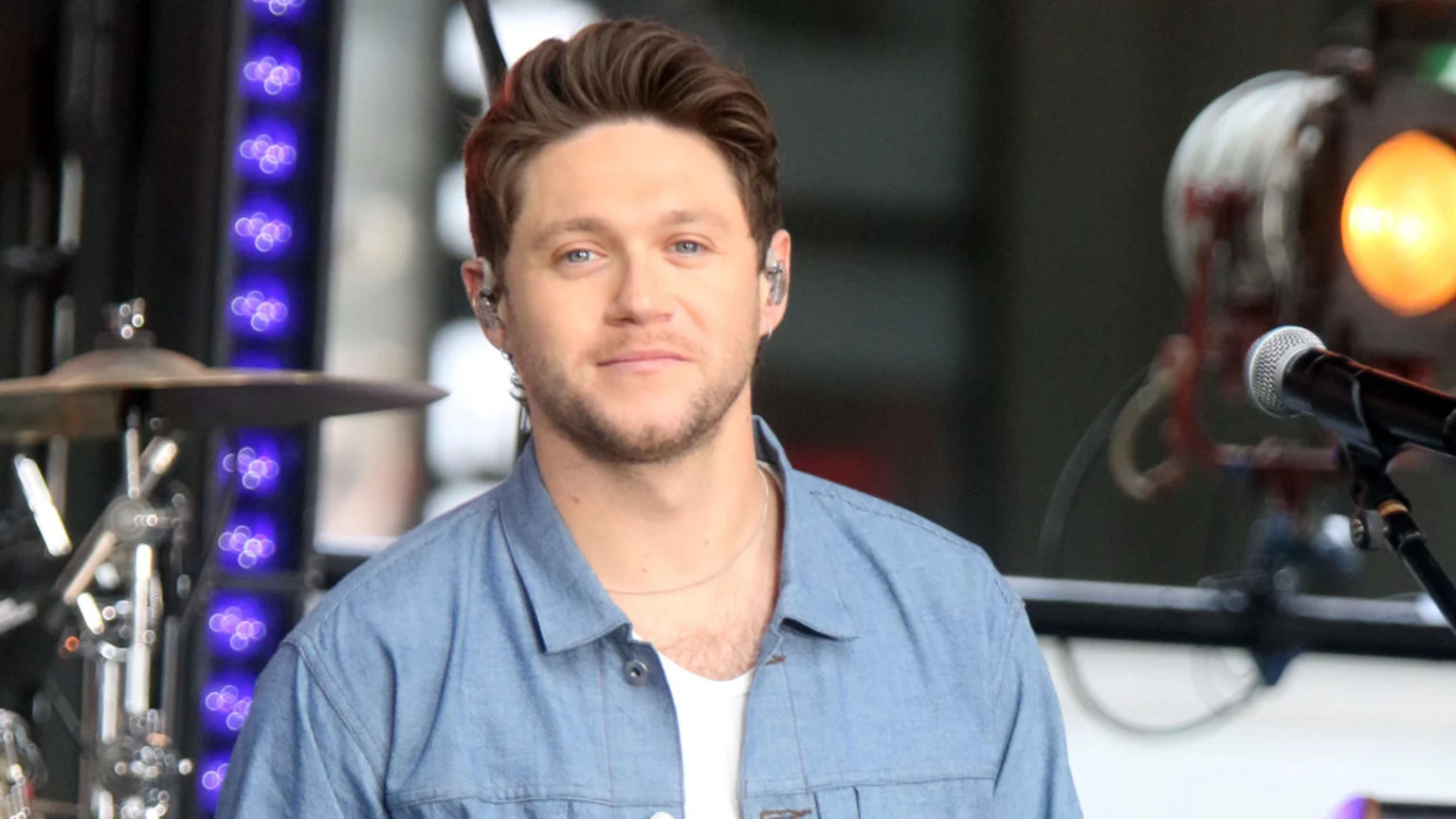 A man with brown hair and a beard, wearing a denim shirt over a white t-shirt and in-ear monitors, smiles slightly while out-of-focus stage equipment is visible in the background.