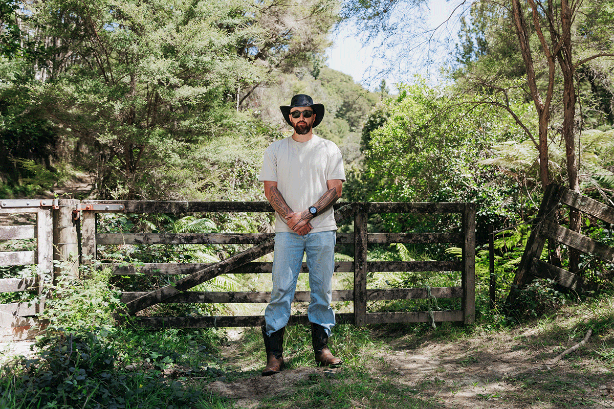 A bearded man in a black hat, sunglasses, light t-shirt, jeans, and cowboy boots stands with crossed arms in front of a rustic wooden fence in a sunny, green forest.