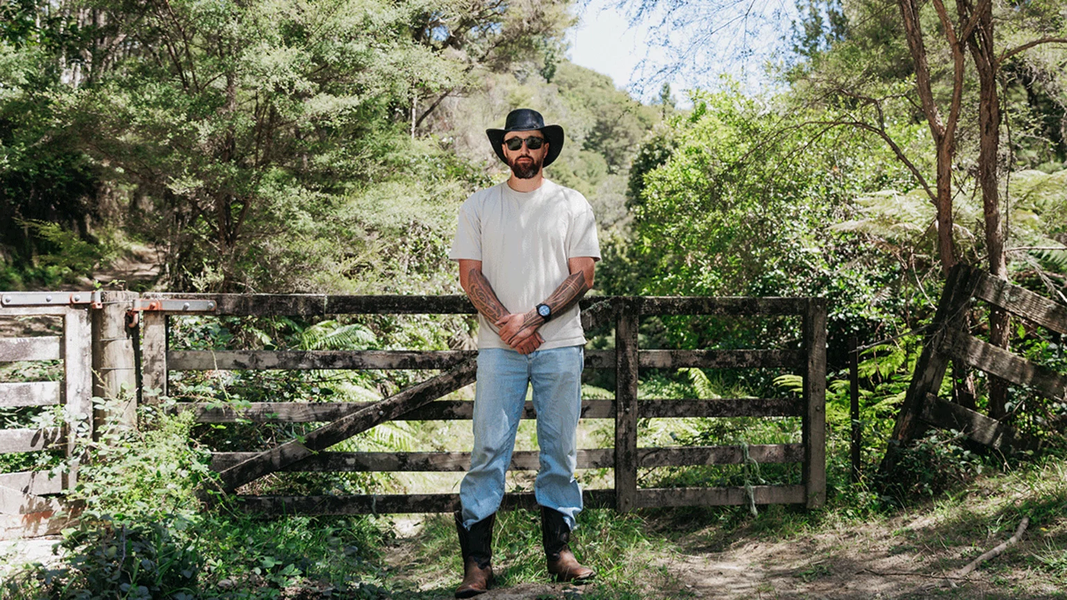 A bearded man in a black hat, sunglasses, light t-shirt, jeans, and cowboy boots stands with crossed arms in front of a rustic wooden fence in a sunny, green forest.