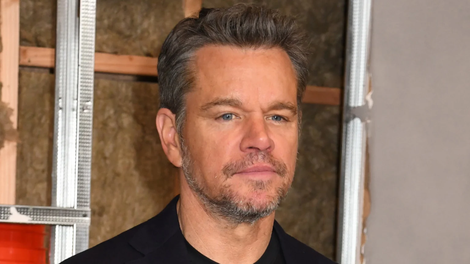 A close-up of actor Matt Damon with salt-and-pepper hair and a beard, wearing a dark jacket, against a background of unfinished construction.