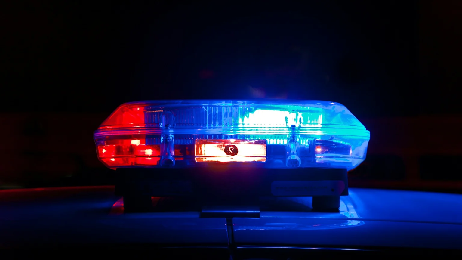 A close-up of a police light bar glowing red and blue on top of a vehicle in a dark setting, with reflections visible on the car's surface.