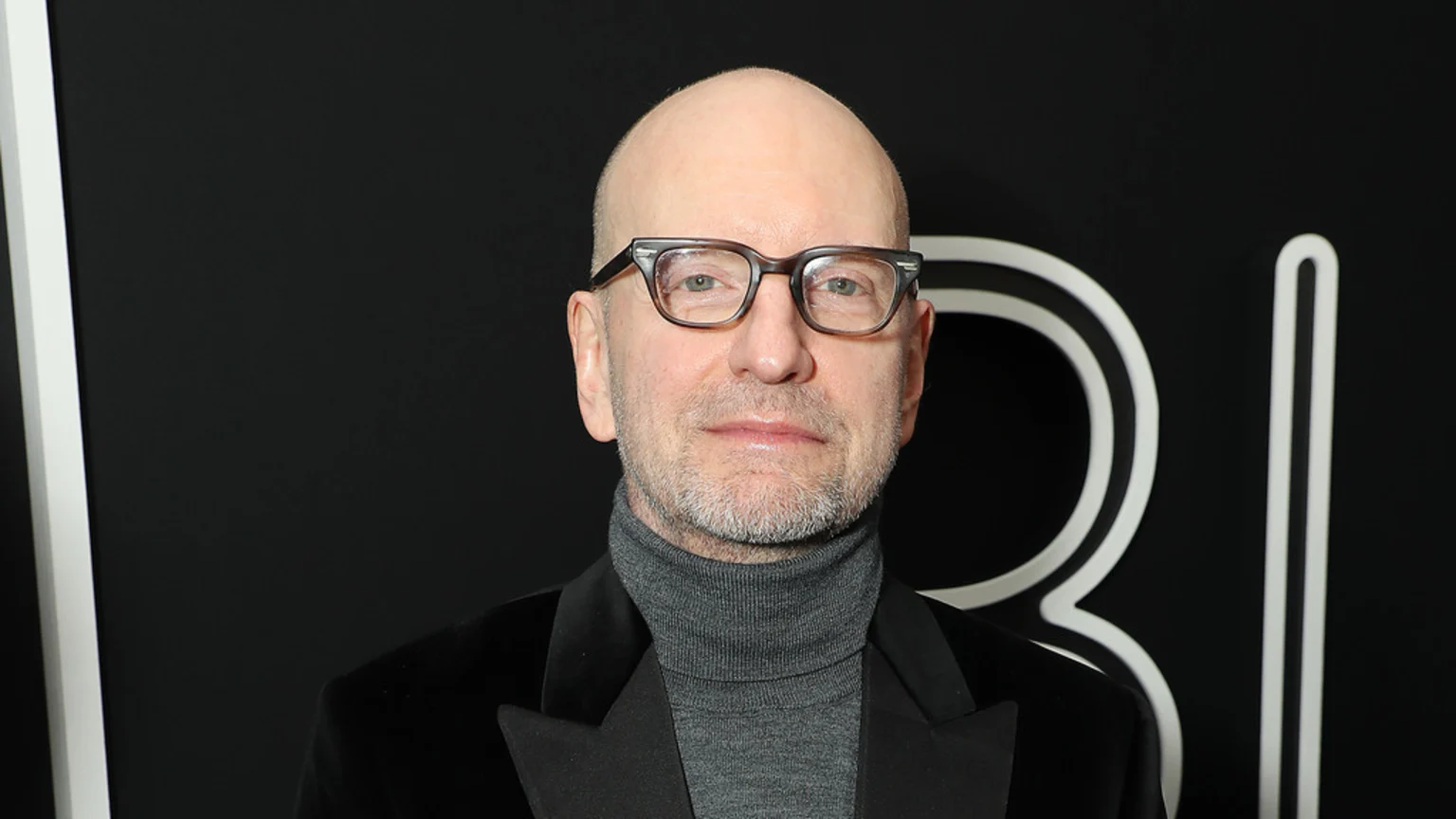 Steven Soderbergh, a bald man with a short beard and glasses, wearing a gray turtleneck and black velvet jacket, looks directly at the camera against a black background with white abstract shapes.