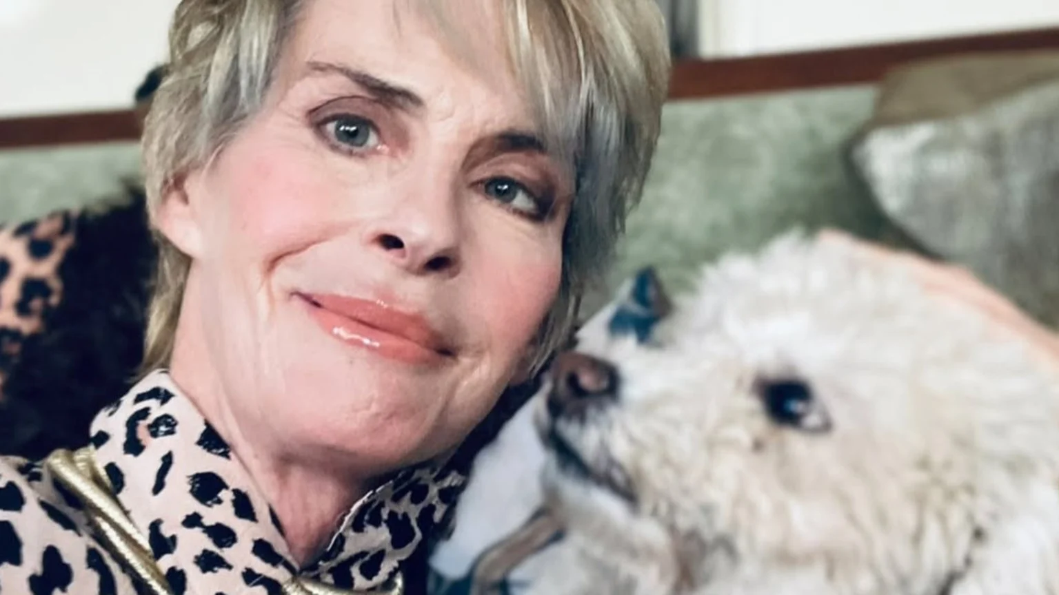 Annabel Schofield with short, light hair, rosy cheeks, and glossy lips, smiles in a close-up selfie, wearing a leopard print top, with a fluffy white dog looking up at her.