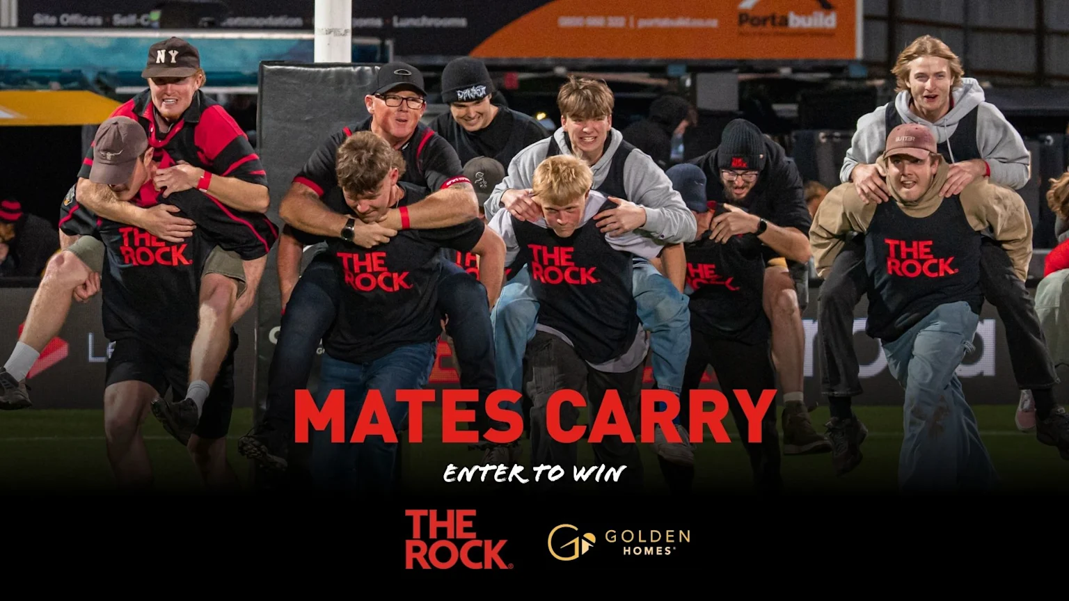 A group of excited men and young adults participate in a "mates carry" competition, with some carrying others piggyback style across a field, all wearing "THE ROCK" shirts.