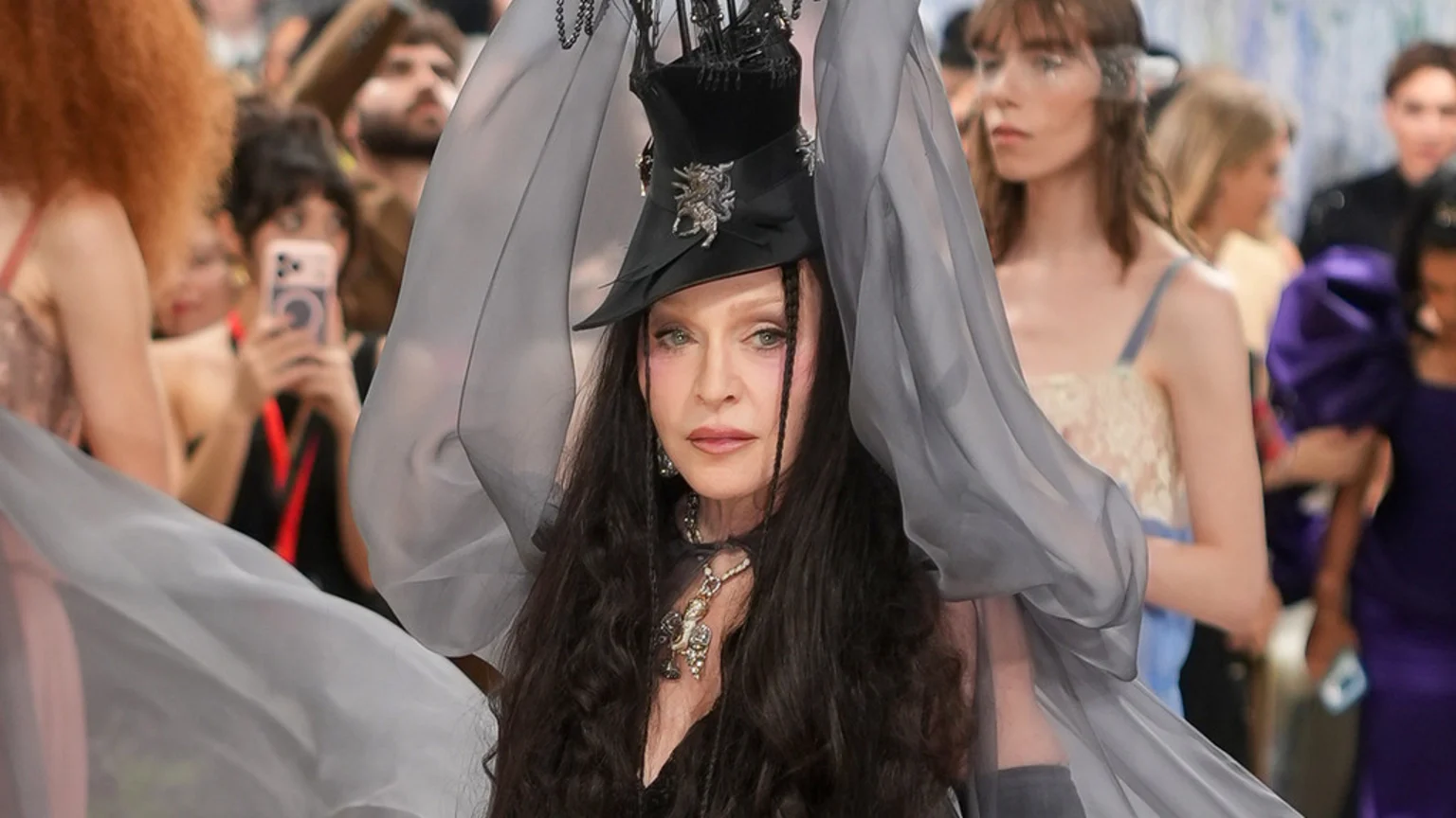 Madonna in an elaborate black hat, dark outfit, and a prominent necklace, holding up flowing sheer grey fabric, with blurred spectators in the background.