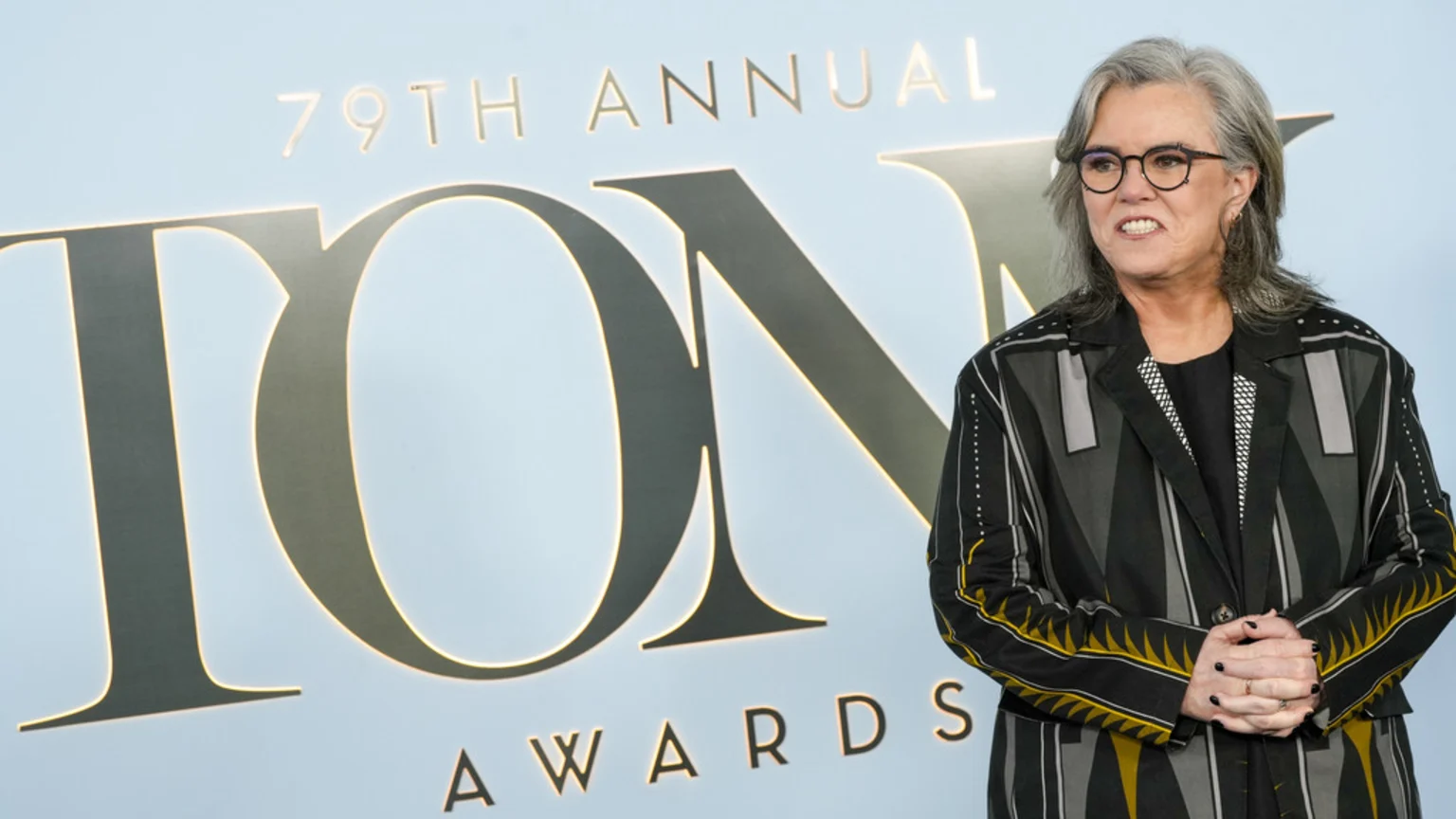 Rosie O'Donnell, a woman with gray hair and glasses, smiles slightly, wearing a patterned blazer in front of a "79th Annual Tony Awards" sign.
