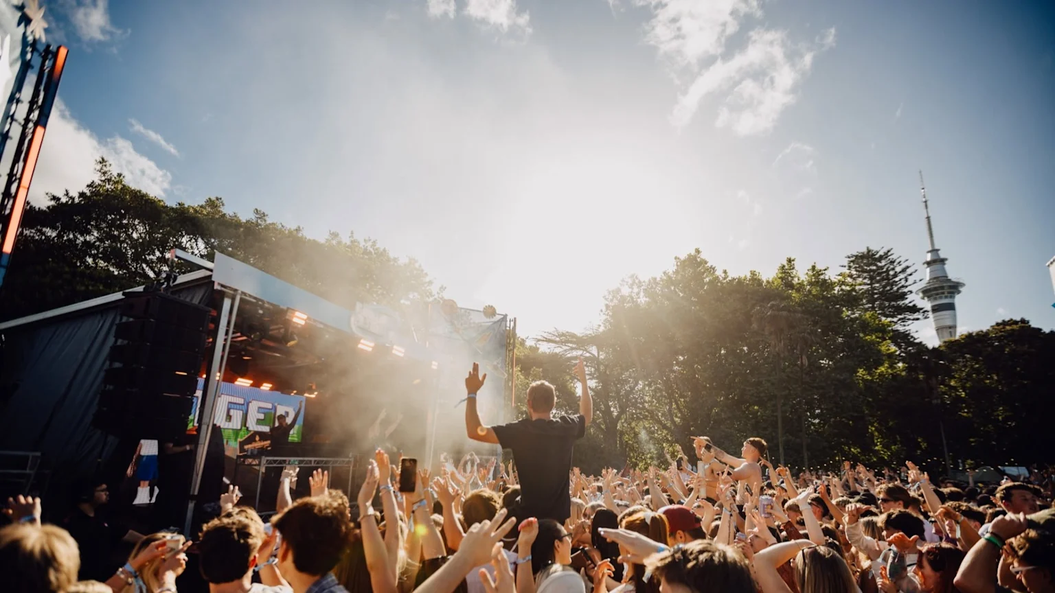 A large crowd with hands raised at an outdoor music festival, with a DJ on stage, trees, and the Sky Tower visible in the background under bright sunlight.