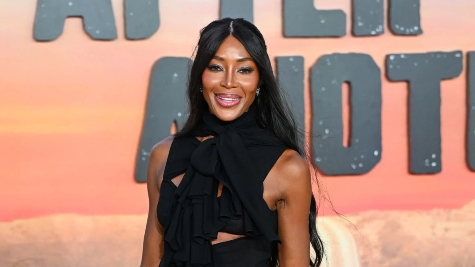 Supermodel Naomi Campbell smiles brightly, wearing a black ruffled top and long dark hair, against a blurred orange-red background with partial grey letters.
