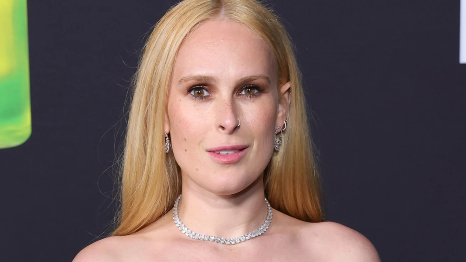 A blonde woman with freckles, light brown eyes, a nose ring, diamond earrings, and a diamond necklace looks forward with a slight smile against a dark background.