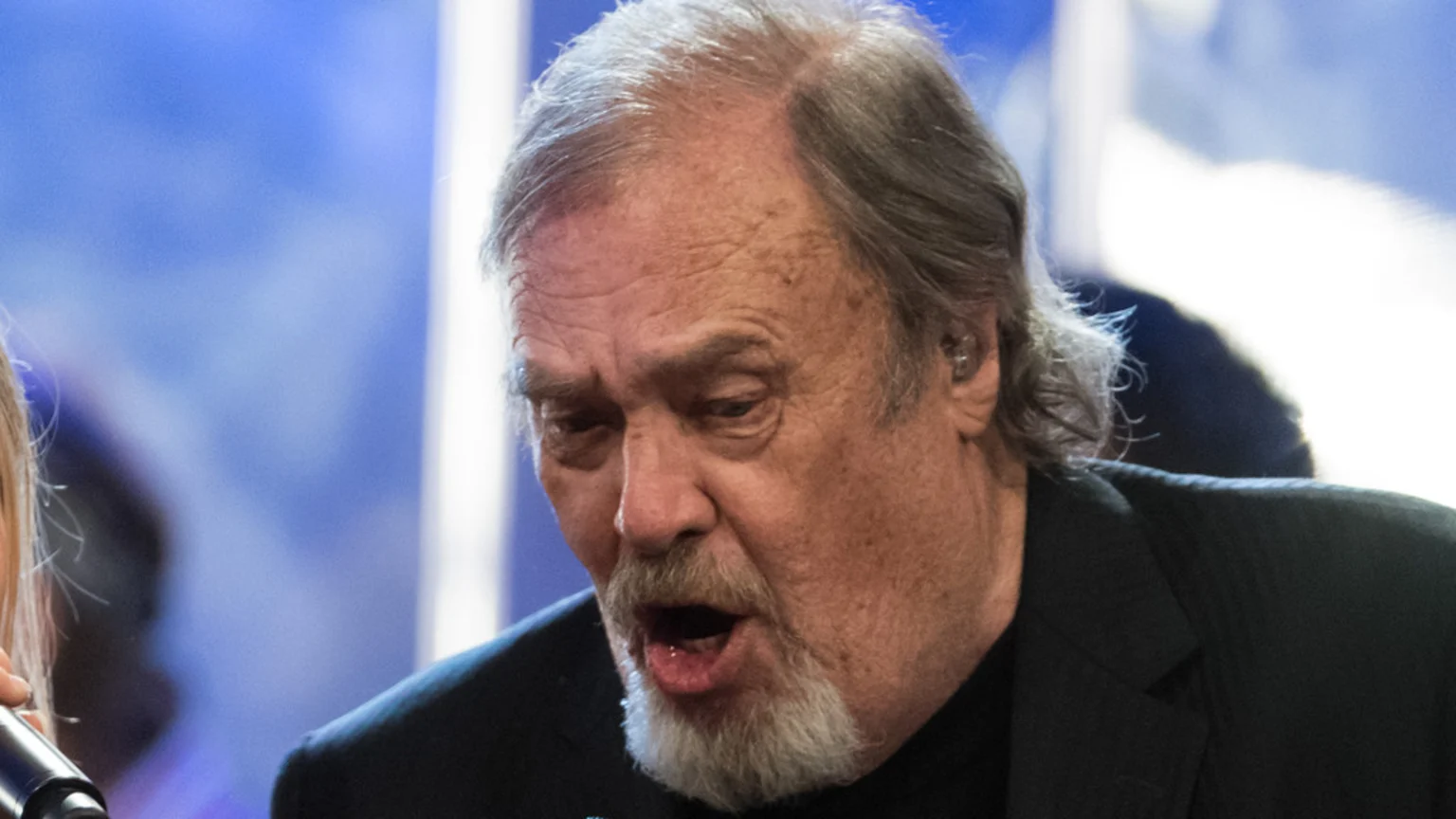 An older man with grey hair and a goatee beard, wearing a dark suit, has his mouth open as if singing, with an earpiece visible in his right ear against a blurred blue background.