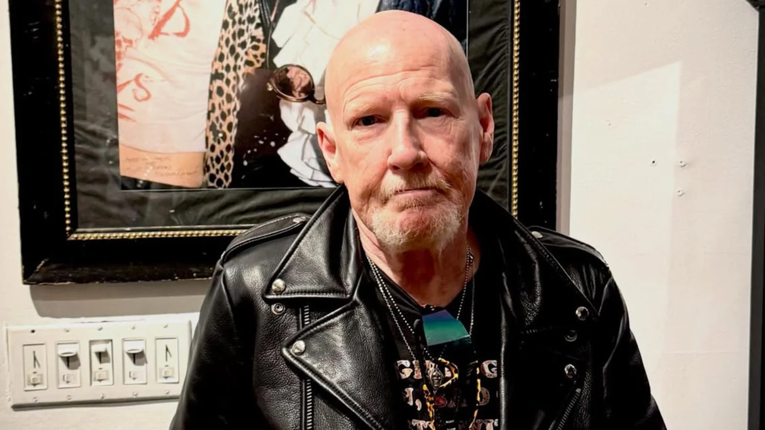 A bald, older man with a goatee and a serious expression wears a black leather jacket and necklaces, standing in front of a framed picture and a light switch panel.