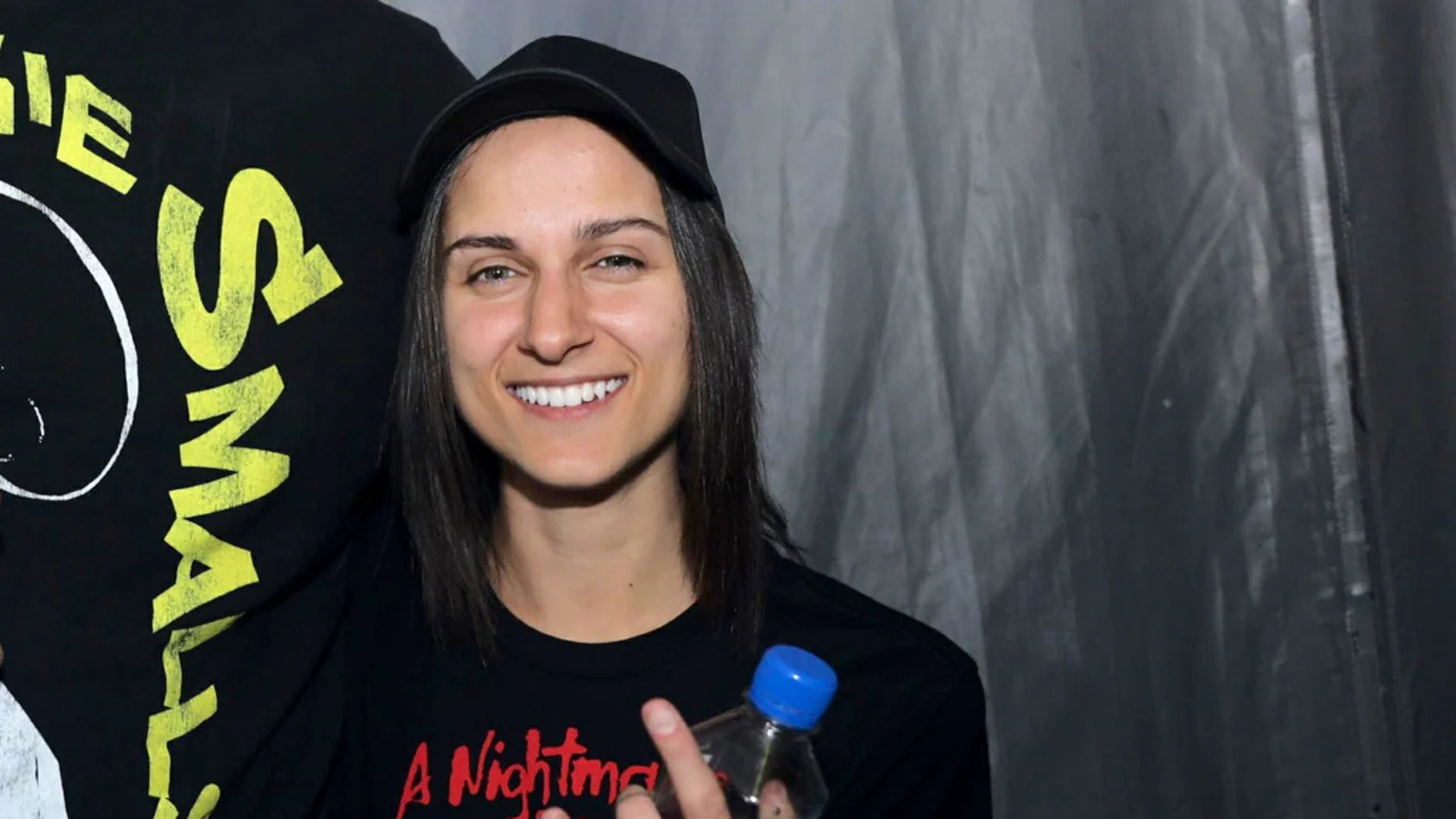 A smiling woman with long dark hair, wearing a black cap and a black t-shirt with red text, looks at the camera while holding a clear bottle with a blue cap. Yellow text on black fabric is visible on her left.
