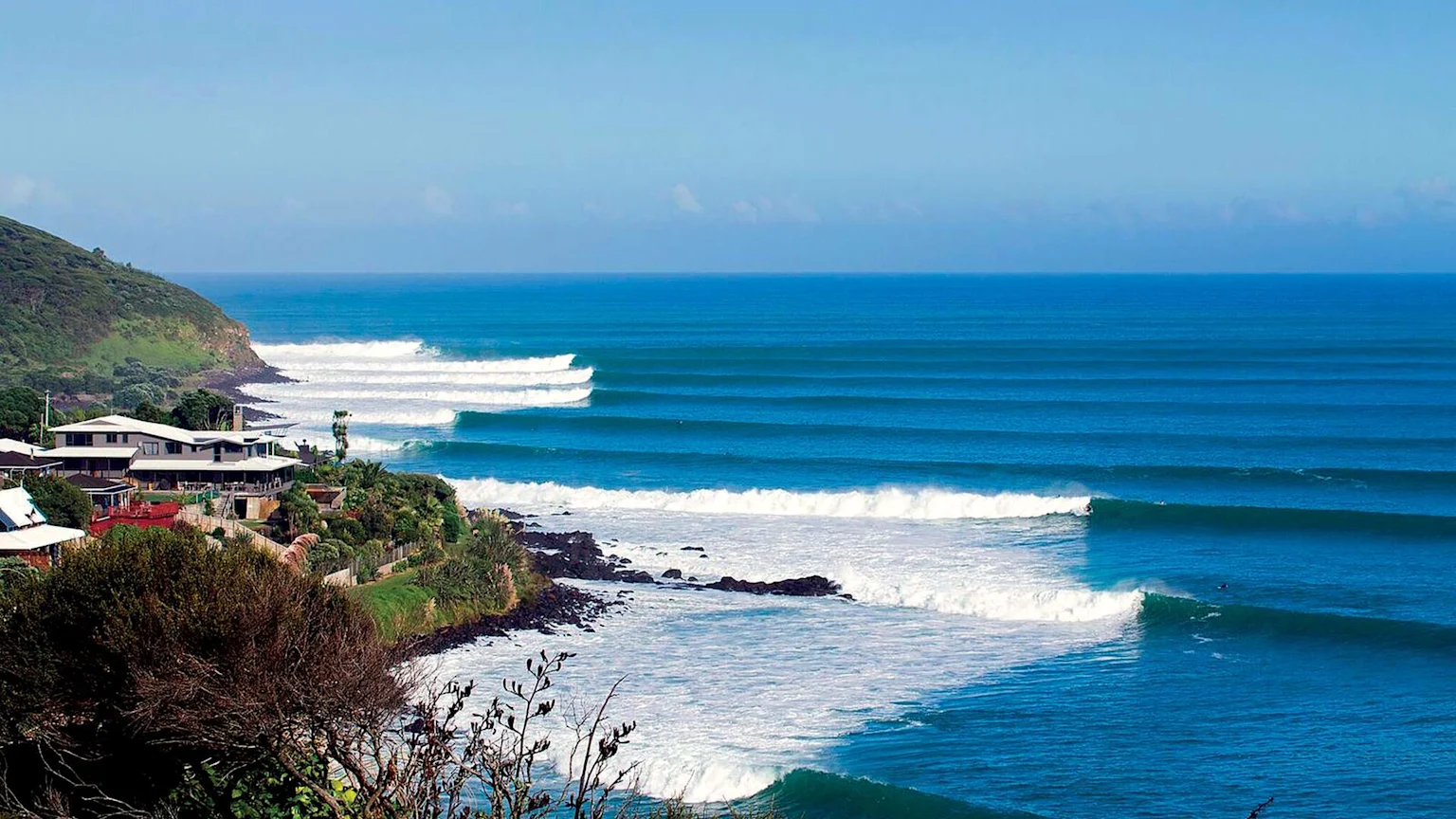 An aerial view of a vibrant blue ocean with perfect lines of waves breaking towards a rocky coast, surfers in the water, and houses nestled on a green hillside under a clear sky.