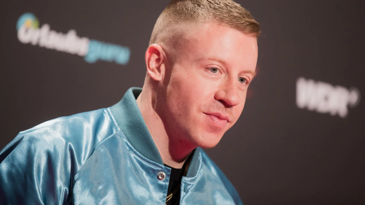 A close-up shows musician Macklemore looking slightly to the right with a faint smile, wearing a shiny light blue satin bomber jacket against a dark background with blurred "Urlaubs guru" and "WDR" logos.