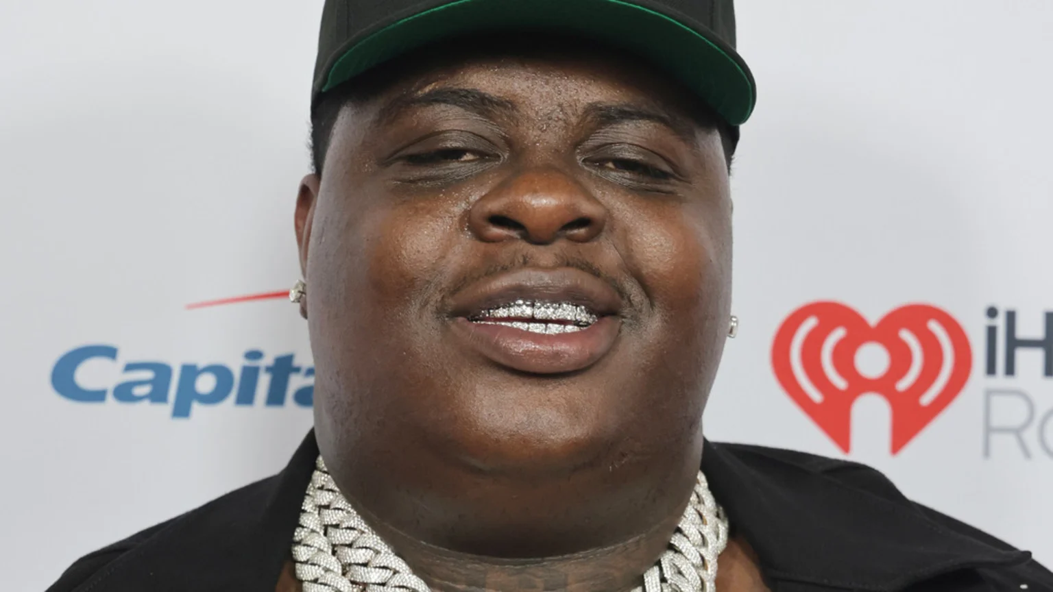 A man wearing a black cap, diamond grills, and jewelry looks at the camera against a white background with "Capital" and "iHeartRadio" logos.
