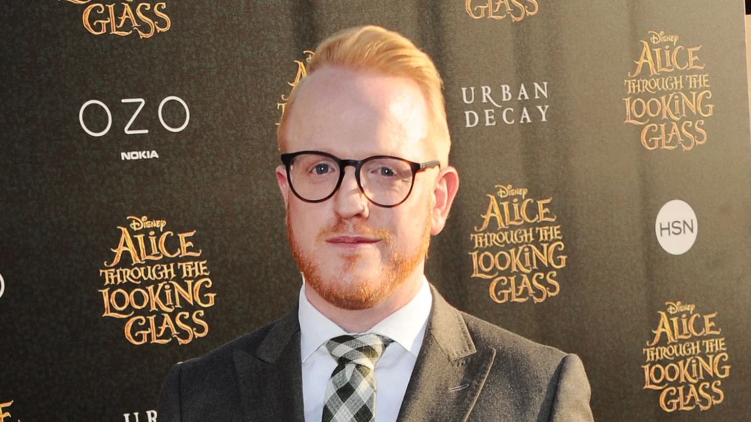 A man with red-blonde hair, a beard, and glasses wears a suit and plaid tie, standing in front of a dark backdrop with "Disney Alice Through The Looking Glass" and other brand logos.