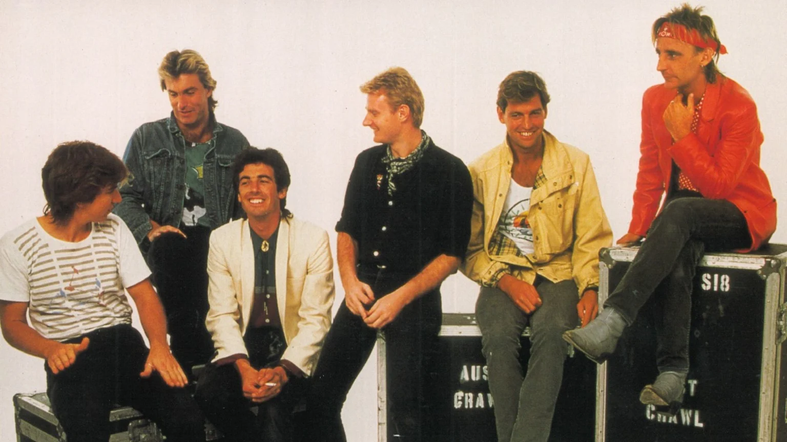 Six men in casual attire are posed, sitting on dark road cases against a light background, some smiling.