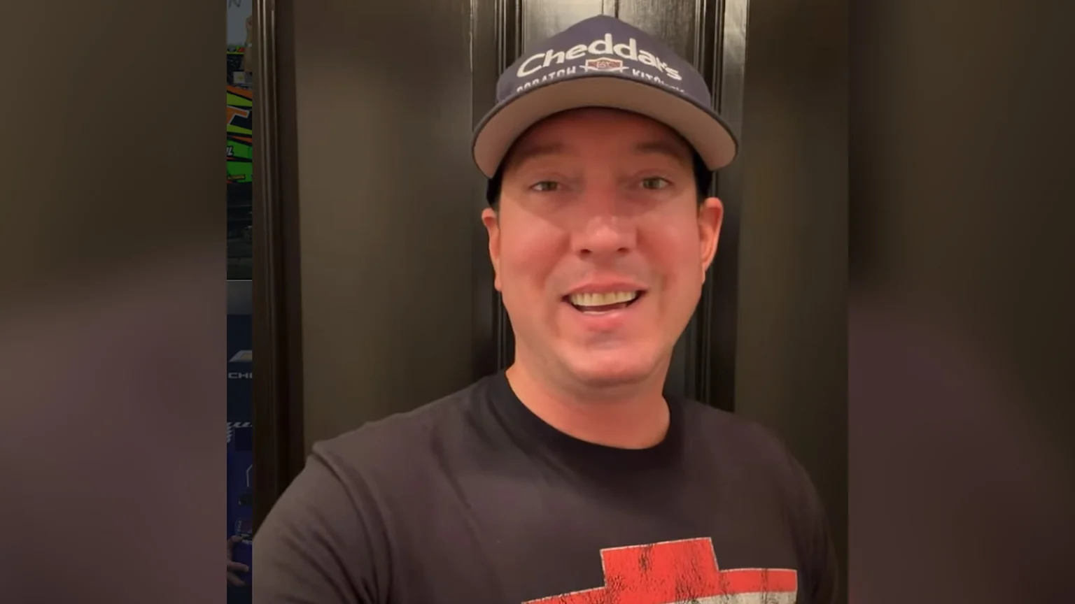 A man wearing a "Cheddar's Scratch Kitchen" cap and a dark t-shirt with a red and white graphic smiles; a racing car livery and "CHI" on a blue background are visible on the left.