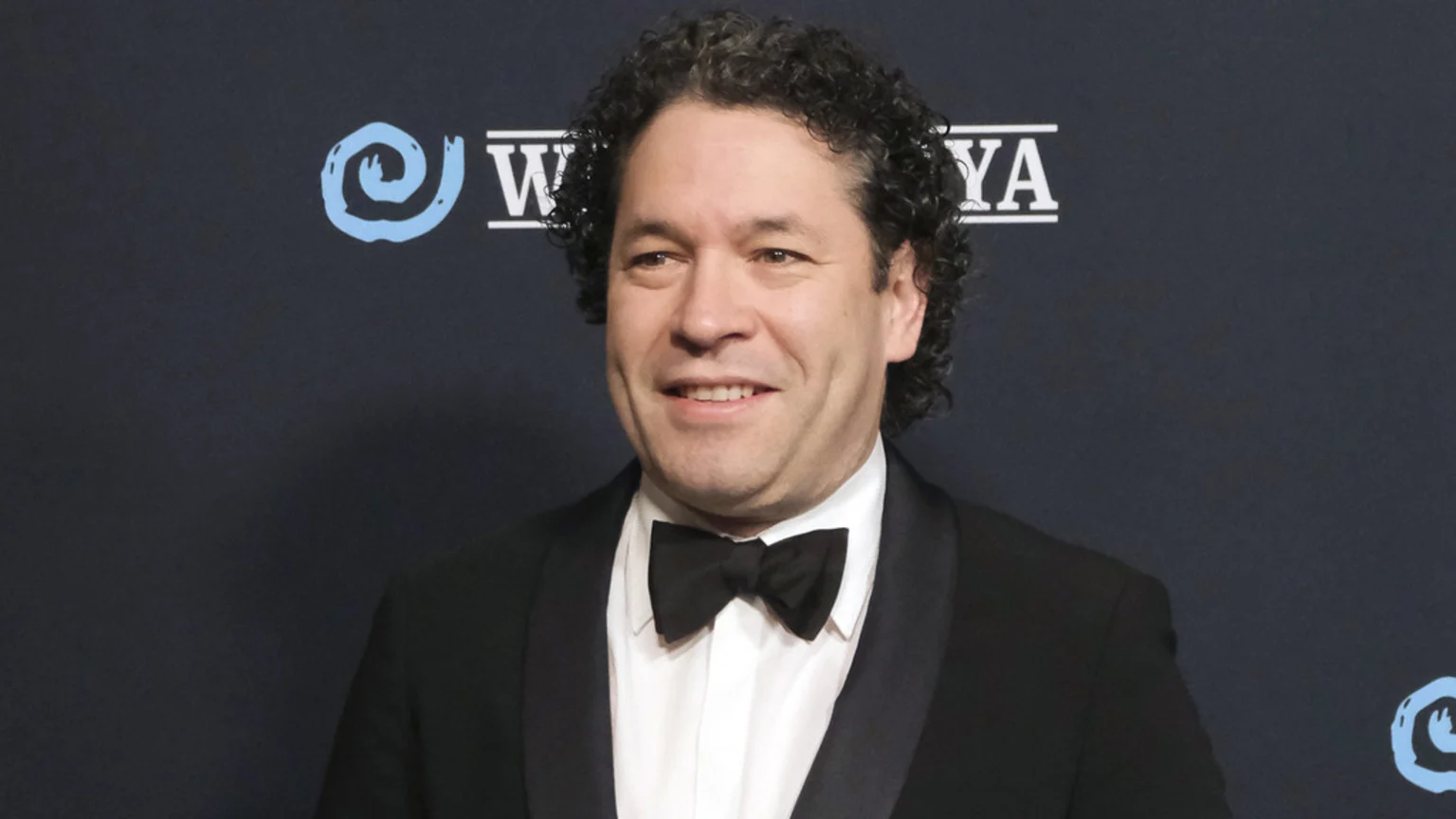 Gustavo Dudamel, a man with curly dark hair, smiles while wearing a tuxedo and black bow tie against a dark background with a blue spiral logo.