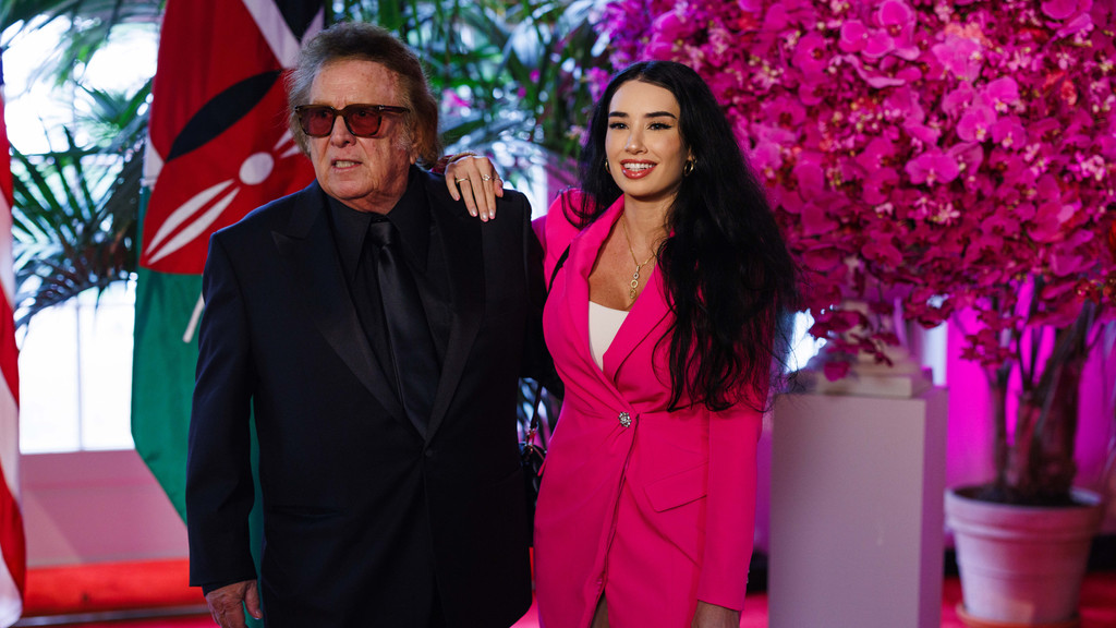 A medium shot indoors shows an older man in a dark suit, black shirt, tie, and orange-tinted sunglasses standing with a younger woman in a bright pink blazer dress, white top, and gold necklace. The woman smiles with her hand on the man's shoulder. The background features a Kenyan flag with a Maasai shield and spears emblem, large pink orchid flowers illuminated with magenta light, green foliage, a red carpet, and a white pedestal in an event space setting.