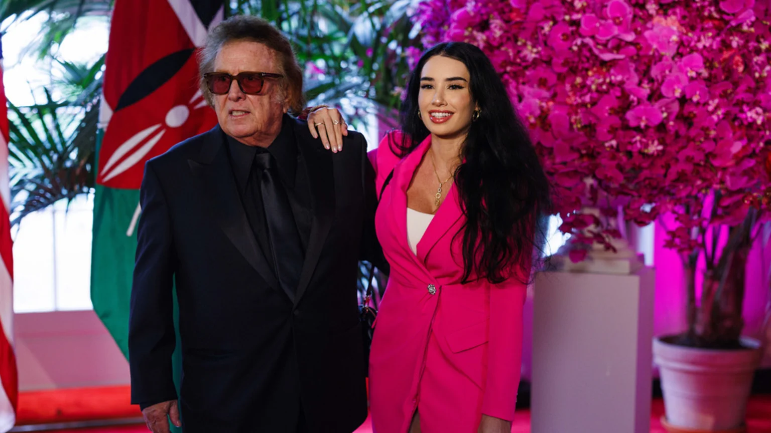 A medium shot indoors shows an older man in a dark suit, black shirt, tie, and orange-tinted sunglasses standing with a younger woman in a bright pink blazer dress, white top, and gold necklace. The woman smiles with her hand on the man's shoulder. The background features a Kenyan flag with a Maasai shield and spears emblem, large pink orchid flowers illuminated with magenta light, green foliage, a red carpet, and a white pedestal in an event space setting.