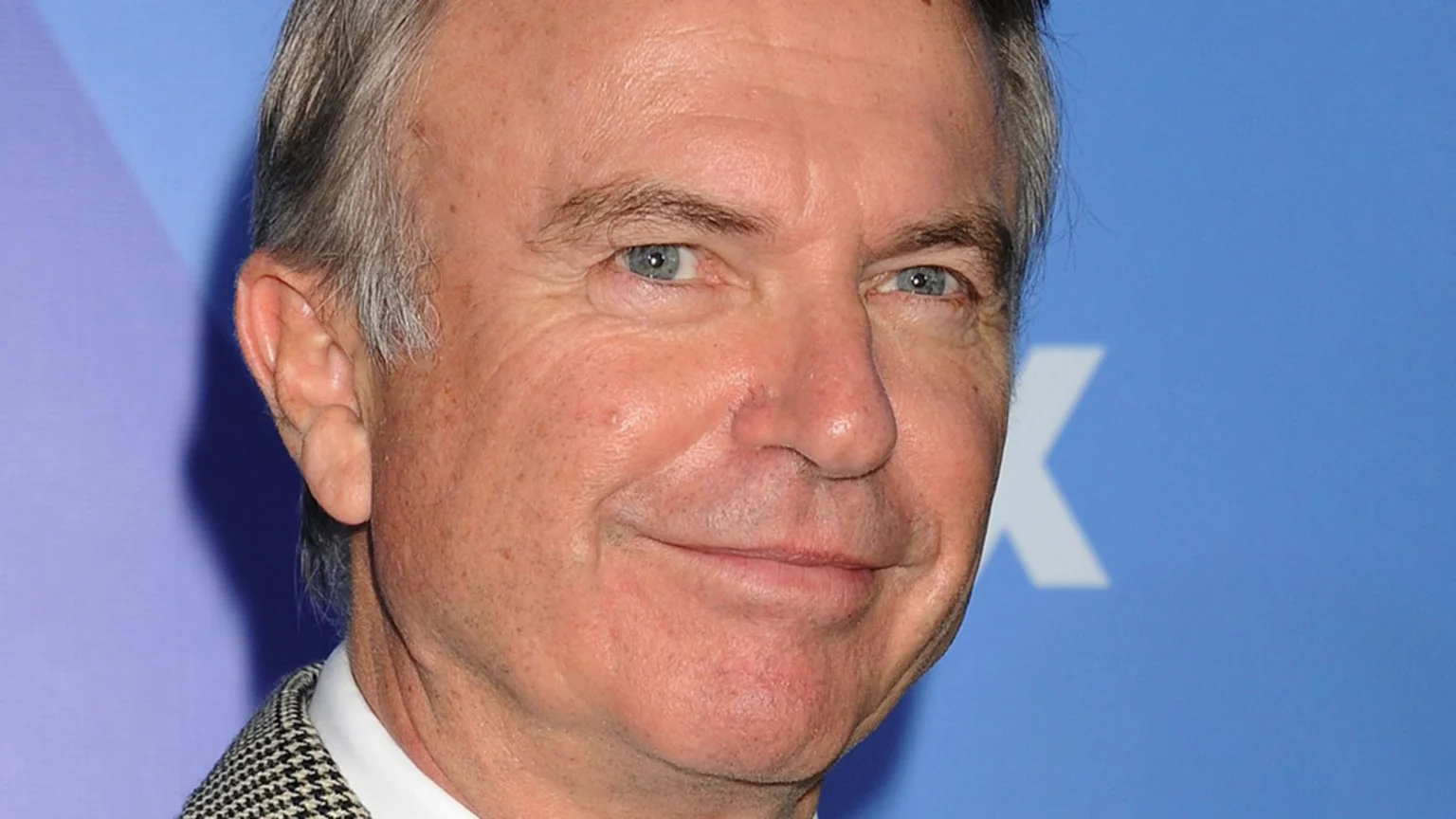Close-up portrait of actor Sam Neill smiling slightly, with graying hair and blue eyes, against a blurred blue background with a white letter K.