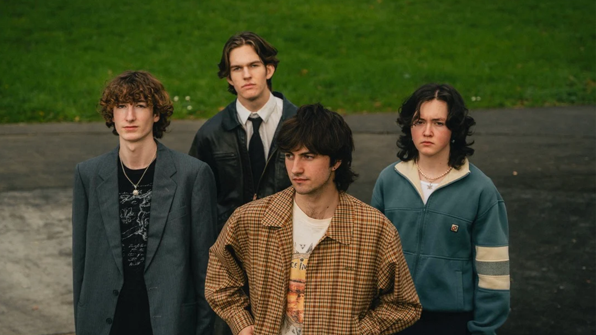 Four young men stand outdoors with serious expressions; one wears a grey jacket, one a leather jacket and tie, one a plaid jacket, and one a teal zip-up jacket.