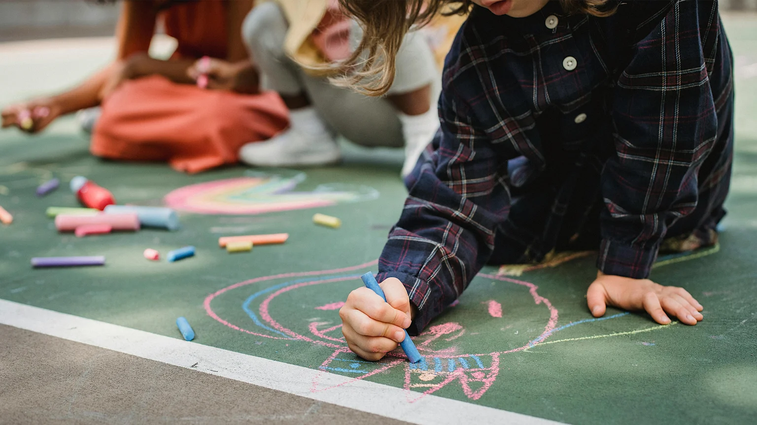 A child in a plaid shirt uses blue chalk to draw on a green paved surface, with other children and colorful chalk scattered around, engaged in outdoor play.