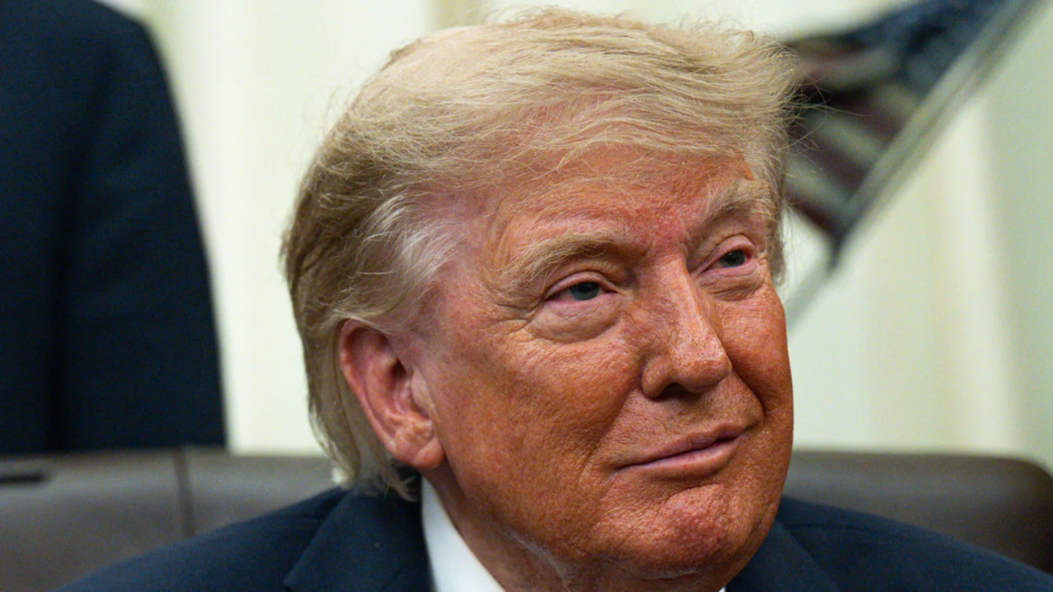 Close-up of Donald Trump, a man with blonde hair and a tanned complexion, wearing a dark suit, smiling slightly and looking to his right.