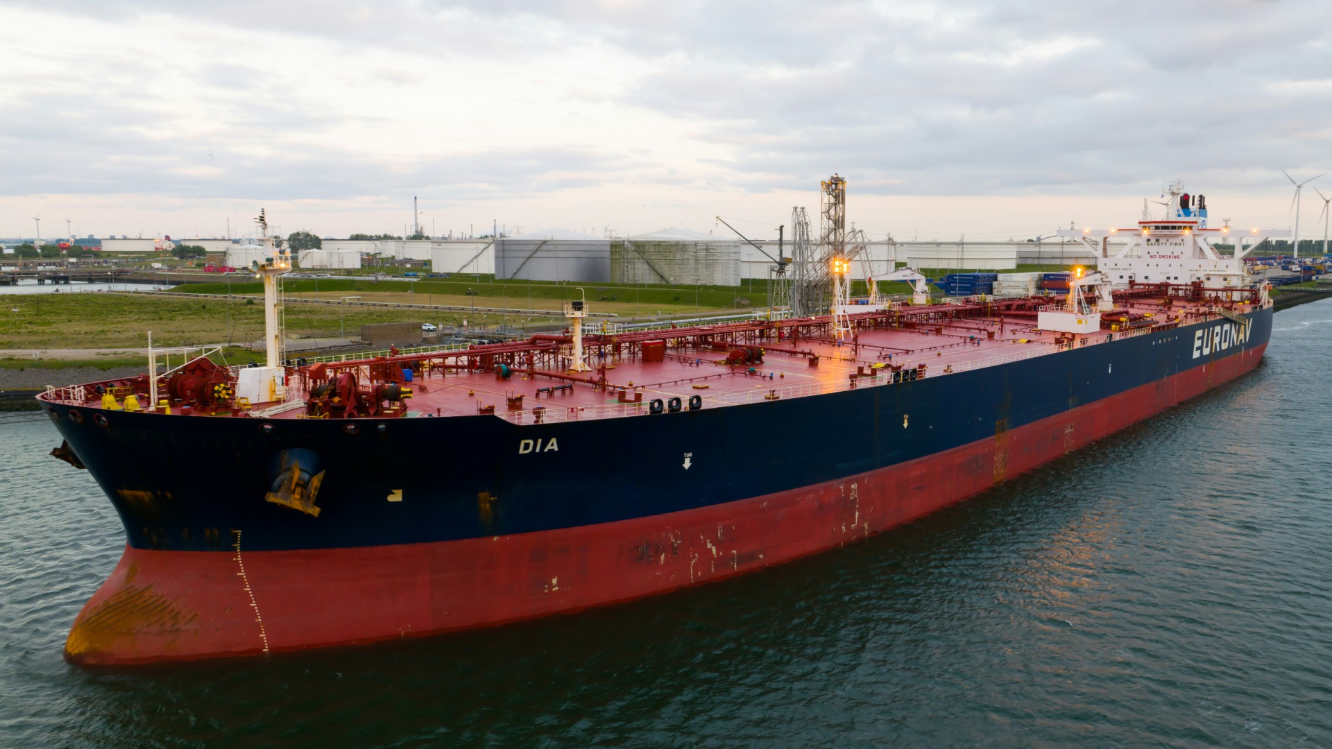 A large oil tanker, named DIA and operated by Euronav, sails in a harbor with industrial facilities and storage tanks visible in the background under a cloudy sky.