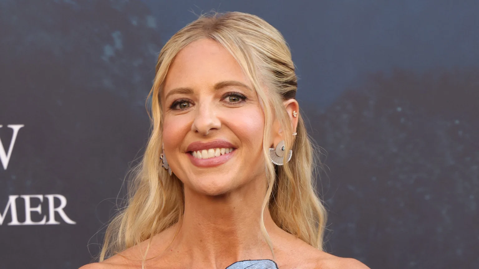 A blonde woman with her hair partly pulled back smiles broadly, showing her teeth, while wearing several earrings and a strapless top.