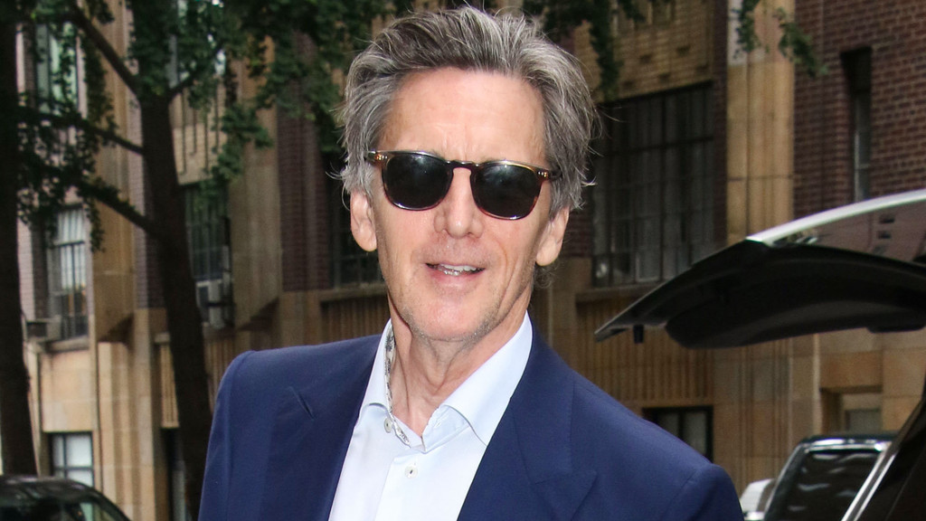 A man with grey hair, wearing sunglasses and a blue suit jacket, smiles at the camera with a blurred background of a building and trees.