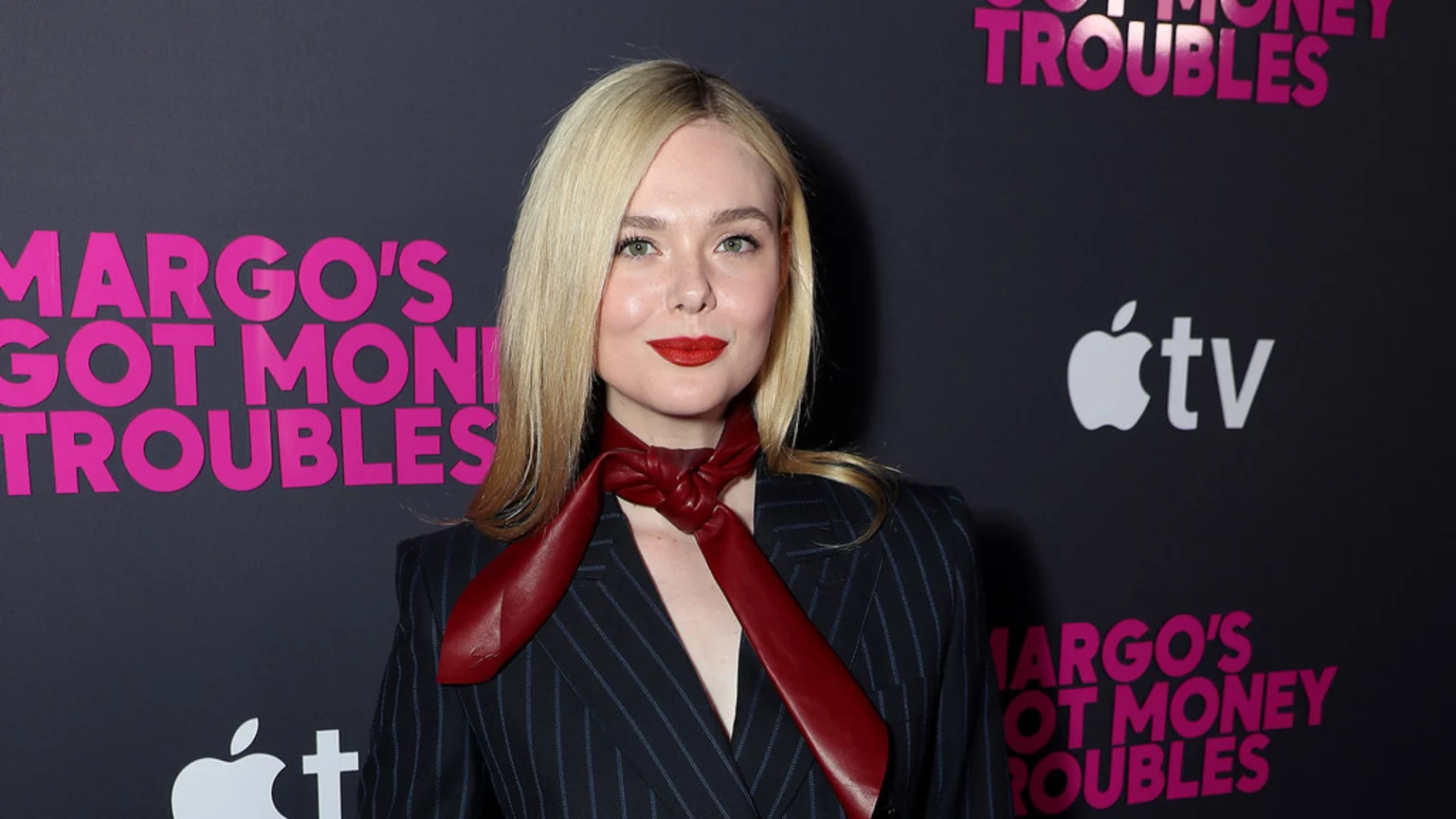 Elle Fanning, a blonde woman with red lipstick, wears a pinstriped blazer and red leather scarf, standing against a dark backdrop with pink "MARGO'S GOT MONEY TROUBLES" text and an Apple TV logo.
