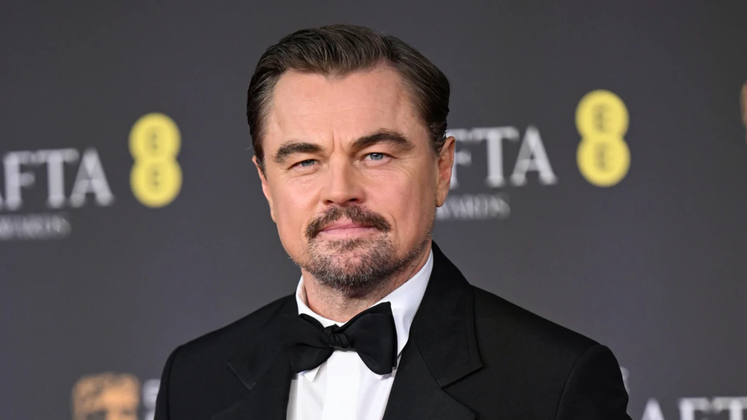 Actor Leonardo DiCaprio in a black tuxedo with a white shirt and bow tie, looking directly at the camera against a blurred dark background with BAFTA logos.