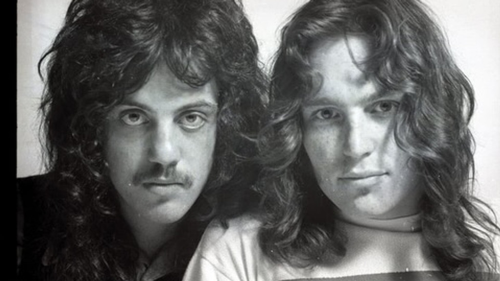 Black and white photo of two men, standing close together, with long, curly hair, one wearing a mustache, looking at the camera.