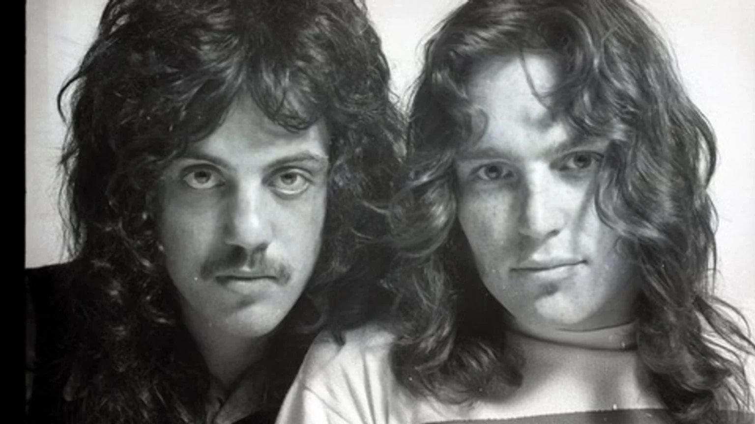 Black and white photo of two men, standing close together, with long, curly hair, one wearing a mustache, looking at the camera.