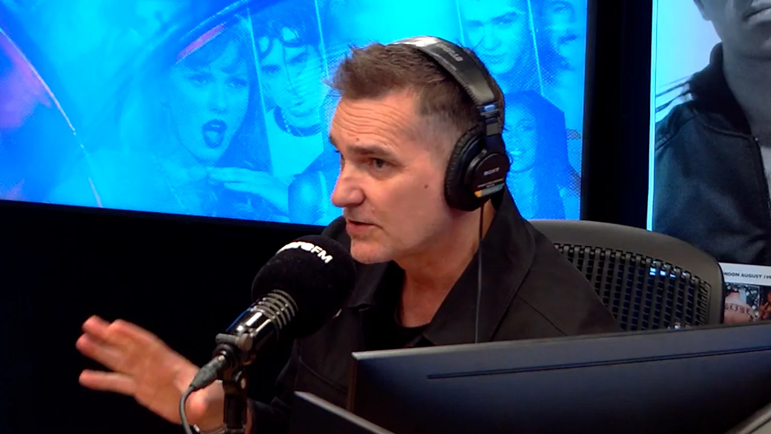 A middle-aged man wearing black Sony over-ear headphones speaks into an RFR FM microphone in a brightly lit radio studio. Behind him, a large blue screen displays stylized images including Taylor Swift singing and a notice reading 'NOON AUGUST 19'.