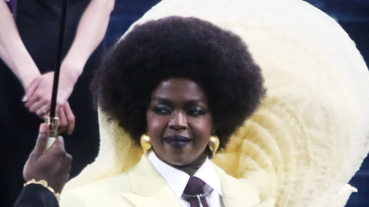 A dark-skinned woman with a large afro, wearing a yellow suit jacket, white shirt, and dark tie, smiles while seated against a large, light-colored, fan-like structure.