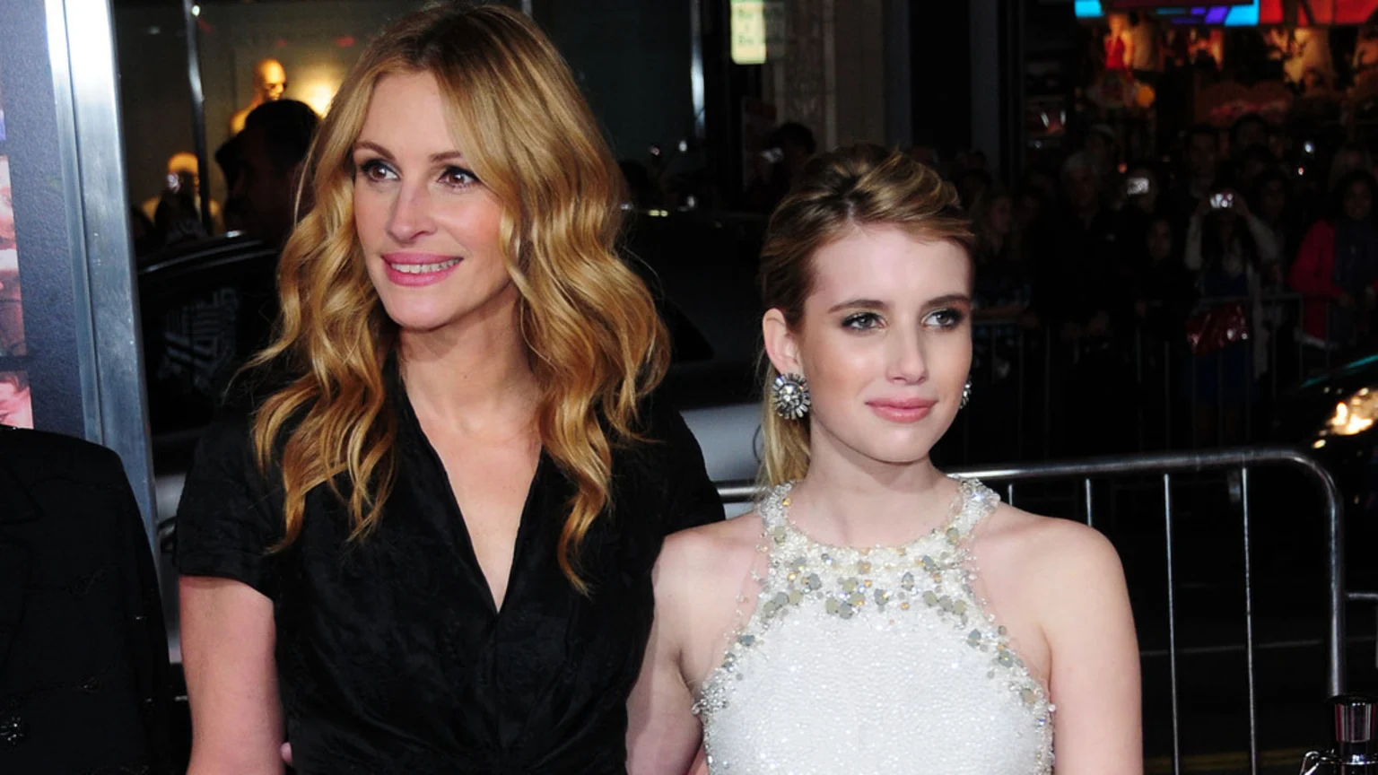 Julia Roberts, with long wavy blonde hair in a black top, and Emma Roberts, in a white sequined dress, pose smiling at an event.