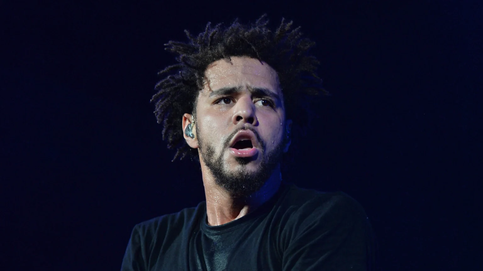 A man with dark dreadlocks and a beard, visibly sweating, wears a dark t-shirt and an in-ear monitor, with his mouth open, looking up and right against a dark background.