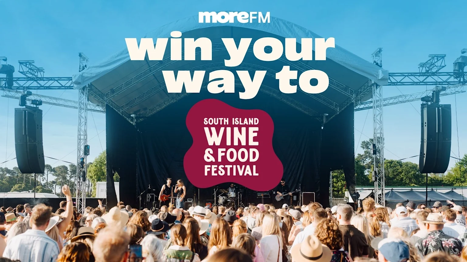 An outdoor music festival scene with a band performing on a stage to a large crowd under a blue sky, featuring text "moreFM win your way to South Island Wine & Food Festival".