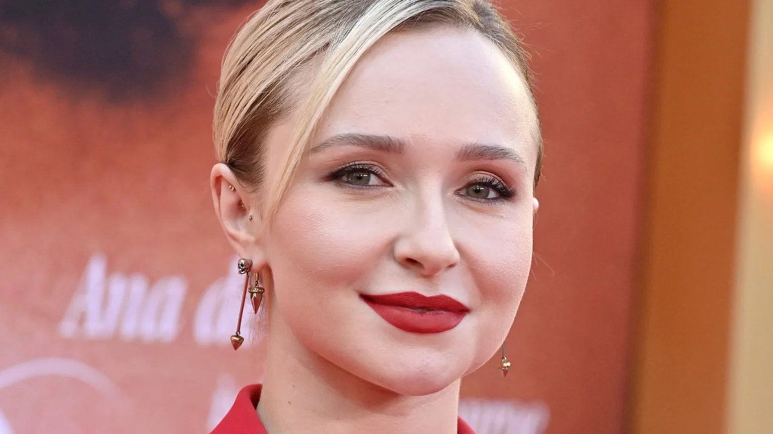 Close-up of Hayden Panettiere with blonde hair, green eyes, and red lipstick, wearing skull dangle earrings and a red top, smiling faintly against a blurred background.