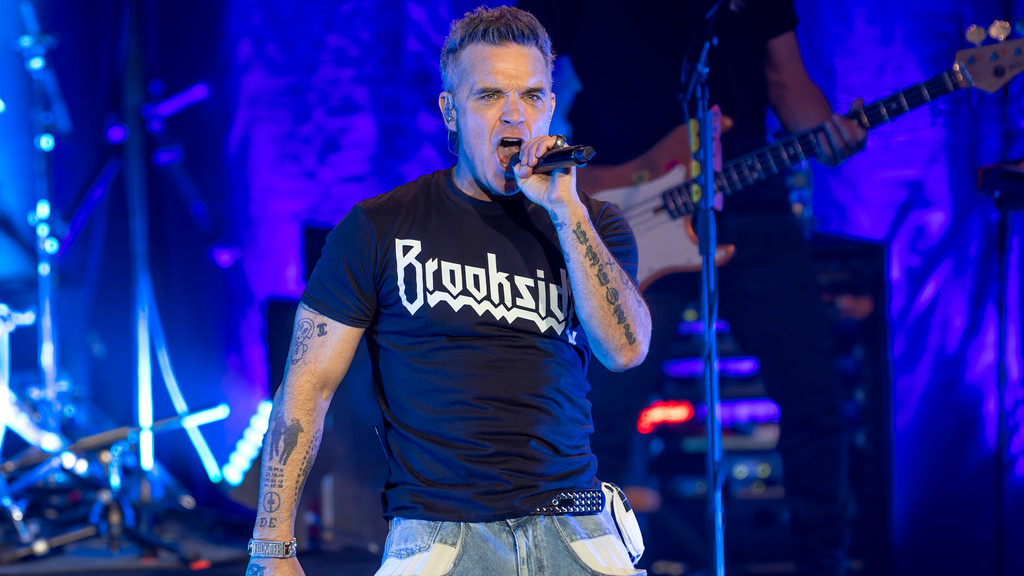 Robbie Williams sings passionately into a microphone on a blue-lit stage, wearing a black "Brookside" t-shirt, jeans, and displaying arm tattoos.