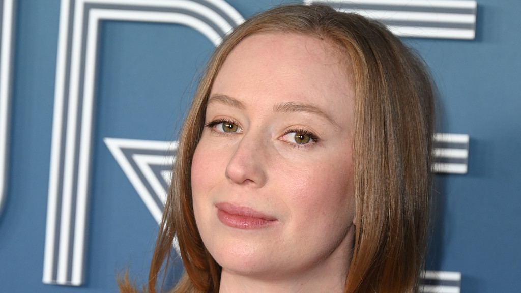 Close-up of actress Hannah Einbinder with light brown hair and a soft smile, against a blue background with white geometric letters.