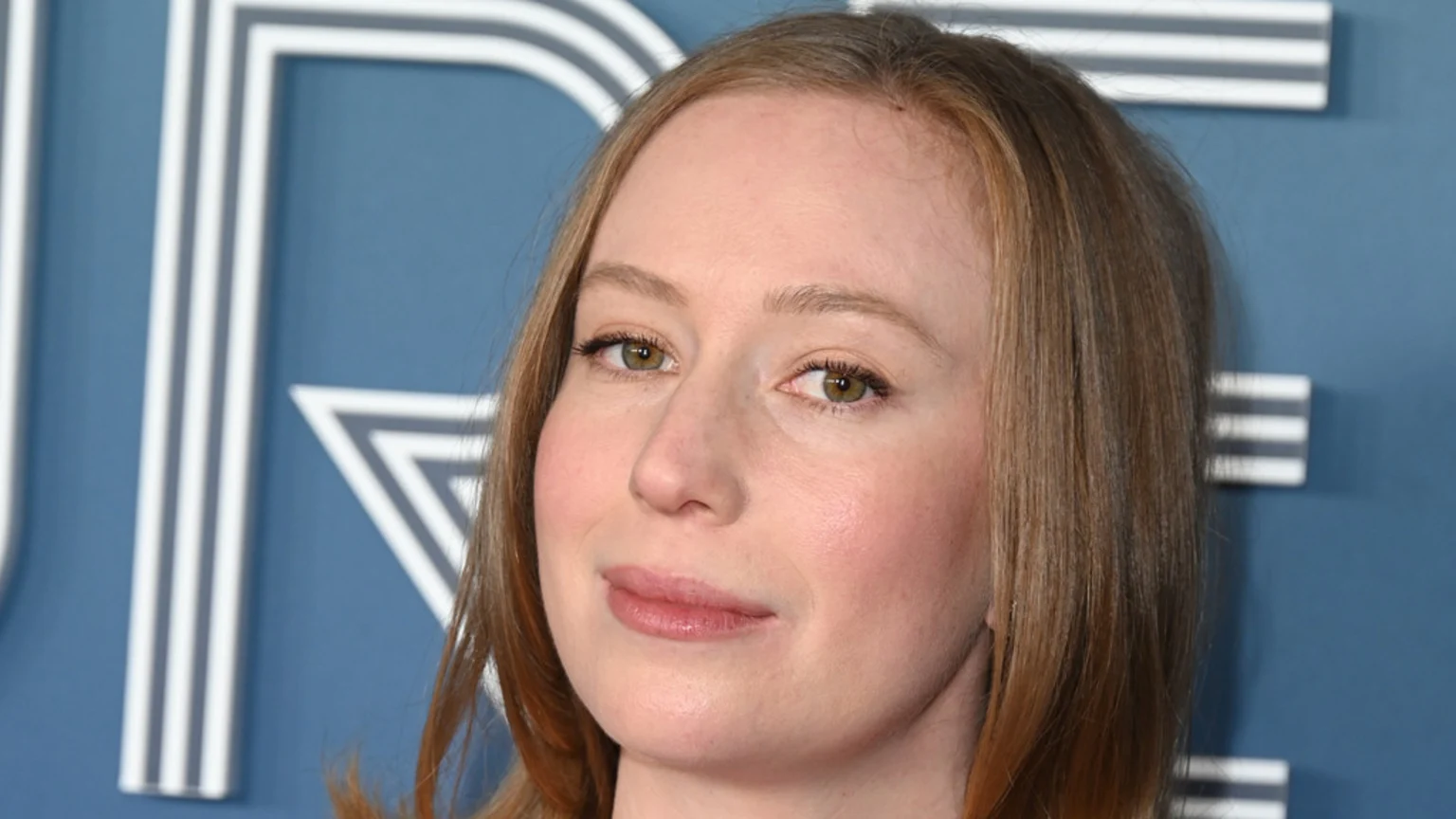 Close-up of actress Hannah Einbinder with light brown hair and a soft smile, against a blue background with white geometric letters.
