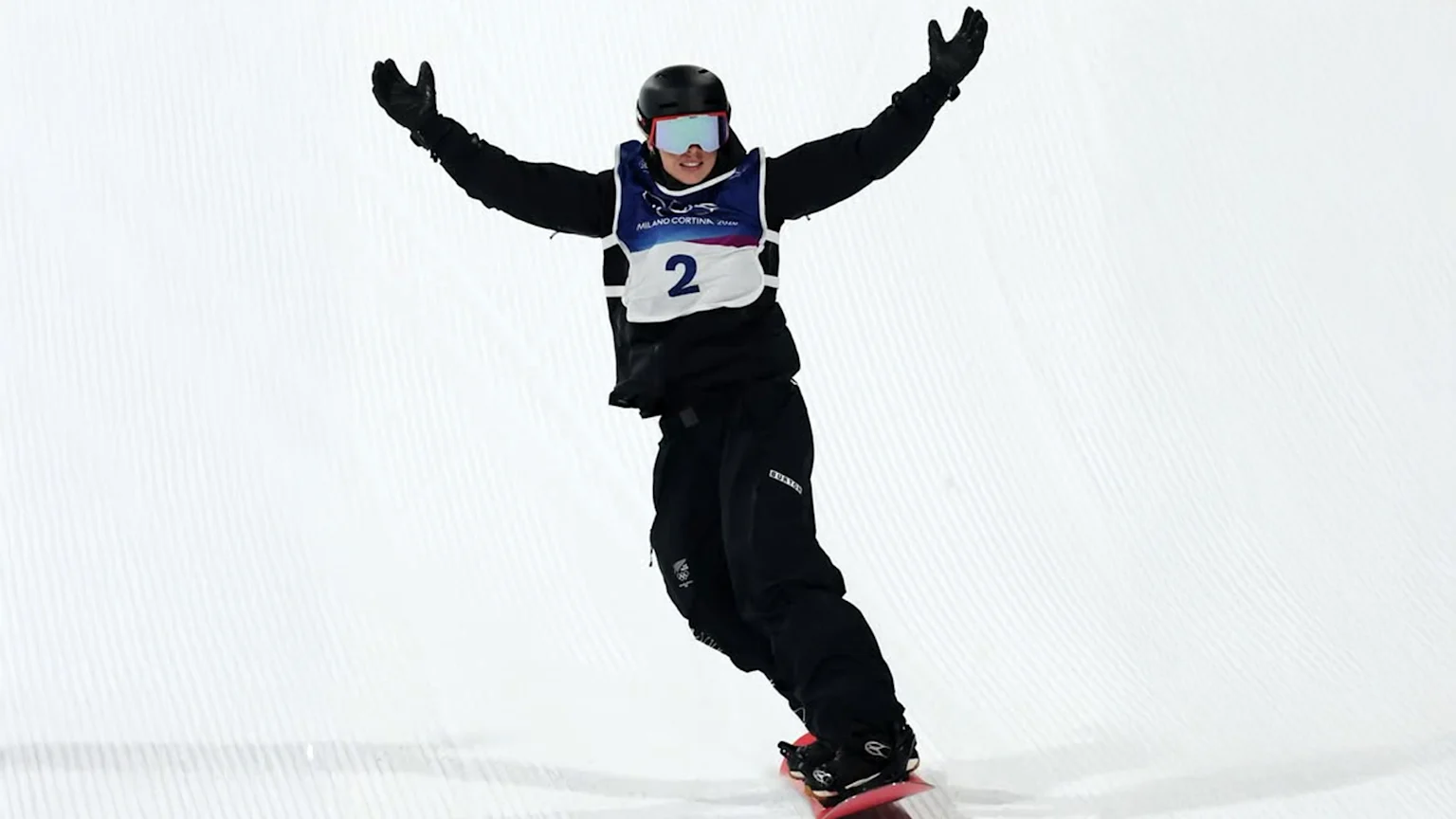 A snowboarder wearing black helmet, blue goggles, and black gear celebrates with arms raised on a groomed snow slope. The white and blue bib numbered '2' shows the event name Milano Cortina 2026 and brand Burton on black pants.